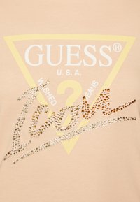 Perzikkleurig T-shirt met een grote gele driehoek en het woord "GUESS" in het wit, versierd met meerkleurige strassstenen.