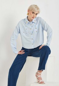 Svetlomodrá košeľa na gombíky s dvoma prednými vreckami a zlatými gombíkmi, v kombinácii s tmavomodrými širokými džínami a bielymi sandálmi na podpätku so remienkami.