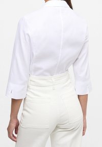 Witte blouse met driekwart mouwen, gemaakt van een textiele stof en een rugnaad, gecombineerd met crème kleurige hoge taillebroek.