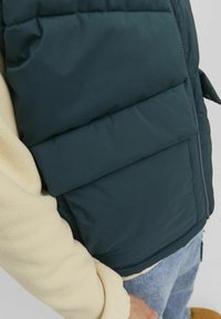 Padded vest in dark green with quilted sections, layered over a beige fleece, featuring a zip closure and side pocket for practicality.