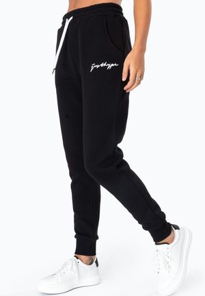 Personne portant un jogger noir avec un cordon blanc et le logo "justhype", associé à des baskets blanches sur fond blanc.