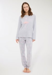 Giovane donna che sta in piedi a piedi nudi, indossando un pigiama grigio chiaro con disegni di ballerine rosa e fiocchi, in posa su uno sfondo bianco semplice.