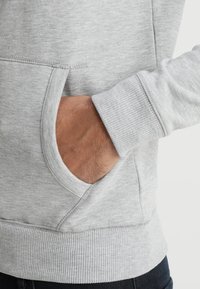 Sweatshirt gris clair avec une poche frontale, des poignets côtelés et une finition texturée. Vue rapprochée de la poche et du détail de la manche.