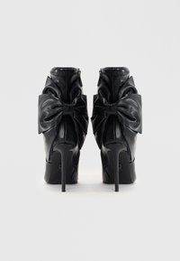 Bottines à talons hauts noires en cuir lisse, avec de grands nœuds à l'arrière et une fermeture éclair élégante.