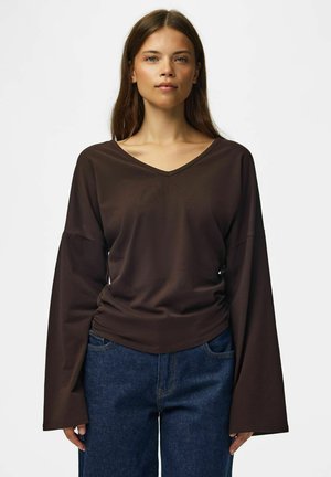 Femme aux cheveux bruns raides porte une chemise marron foncé à manches longues et col en V avec des manches larges et un jean bleu, debout sur fond blanc.