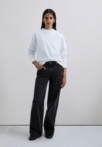 Woman standing with one hand in pocket wearing a white long-sleeve shirt, black wide-leg jeans, and black shoes against a plain background.