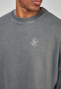 Sudadera gris con cuello redondo acanalado y un pequeño logo bordado intrincado en el lado izquierdo del pecho. Tejido suave y texturizado.