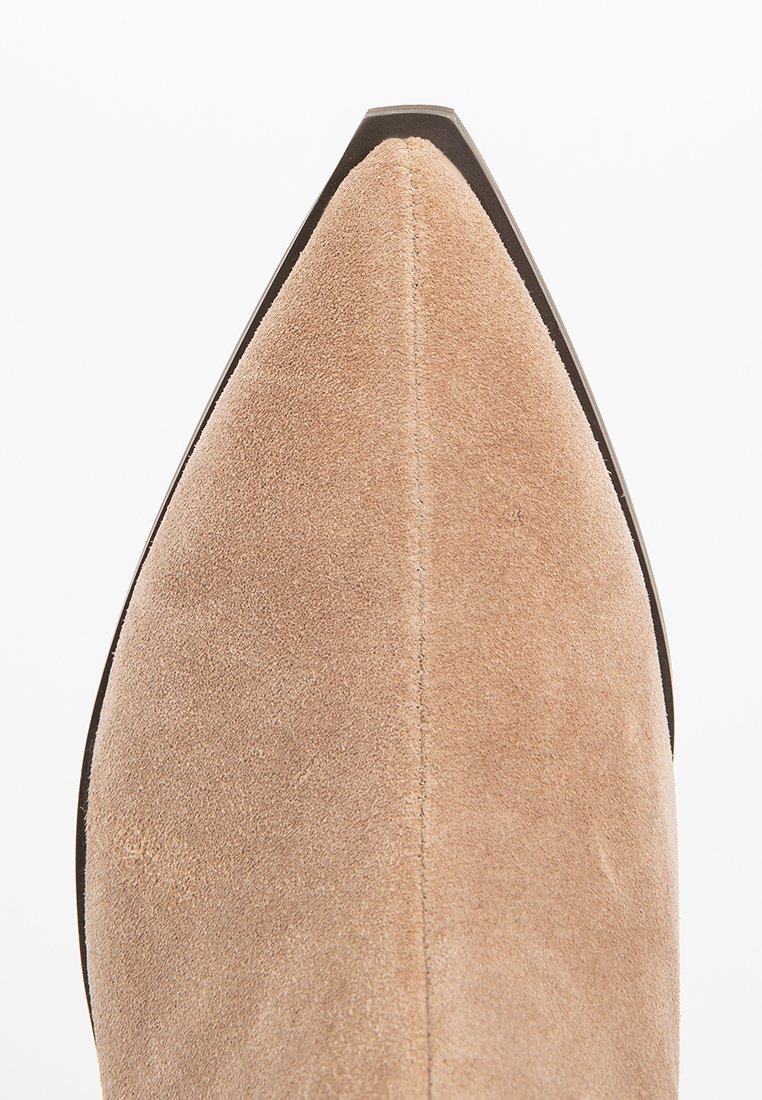 Punta afilada de un zapato de ante beige con costura central visible y ribete oscuro alrededor de la suela, contra un fondo blanco.