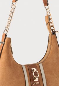 Tasche aus beigem Wildleder mit goldener Kettenriemen, Reißverschlussverschluss und goldenem Guess-Logo auf einem vertikalen Lederriemen auf der Vorderseite.