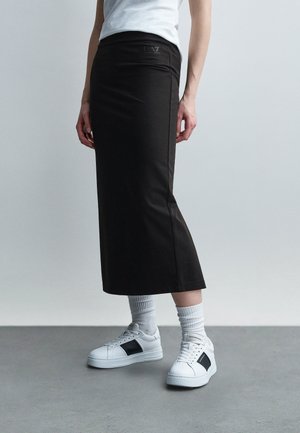 Osoba oblečená v čiernej dlhej sukni, bielych ponožkách, bielych teniskách s čiernymi detailmi a bielom tričku na jednoliatom sivom pozadí.