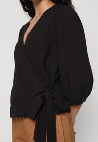 Blusa avvolgente nera con texture, scollatura profonda a V, maniche a sbuffo e dettaglio con nodo laterale, abbinata a pantaloni beige a vita alta.