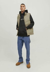 Padded olive vest over a black hoodie, paired with light blue jeans and brown boots. The vest features a zip front and no sleeves.