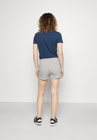 Kvinna med lockigt hår står vänd mot en tom vägg, klädd i en marinblå t-shirt, gråa shorts och svarta Adidas-sneakers med pinka accenter.