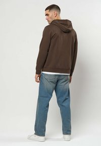 Homme portant un sweat à capuche marron, un jean bleu clair et des baskets blanches, debout dos à un fond uni clair.