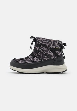 Bota de invierno hasta el tobillo con estampado de leopardo en negro y gris, suela blanca gruesa, puntera negra y cordones ajustables sobre fondo blanco.