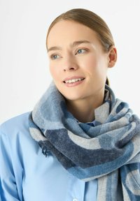 Sciarpa a motivi blu e grigi realizzata in morbida lana, con disegni circolari e bordi frangiati, indossata sopra una camicia blu chiaro.