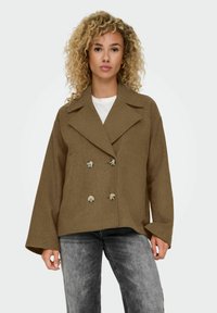 Braune doppelt-knopfige Jacke mit großen Revers und strukturiertem Stoff, ausgestattet mit zwei silbernen Knöpfen und breiten Ärmeln, kombiniert mit einem weißen Oberteil.