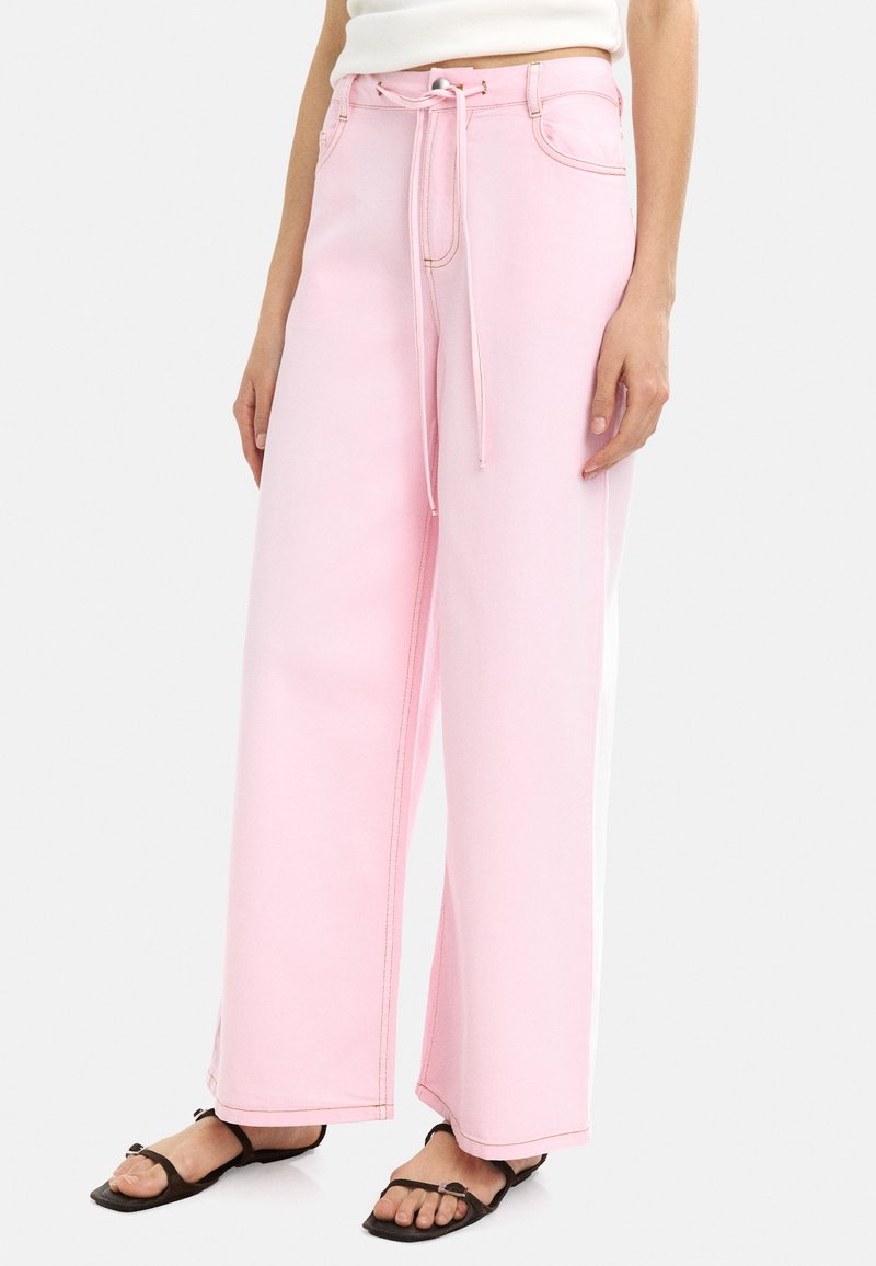 Jasnoróżowe spodnie typu wide-leg wykonane z dżinsu z elastycznym paskiem w pasie. Posiadają kontrastowe przeszycia i boczne paski. W komplecie płaskie przednie kieszenie.