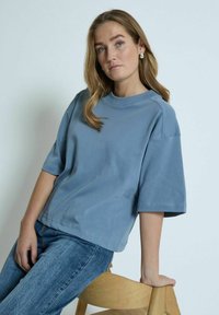 Blå bomulds oversized t-shirt med korte ærmer og rund halsudskæring, parret med lyseblå denim jeans. Træstol i baggrunden.
