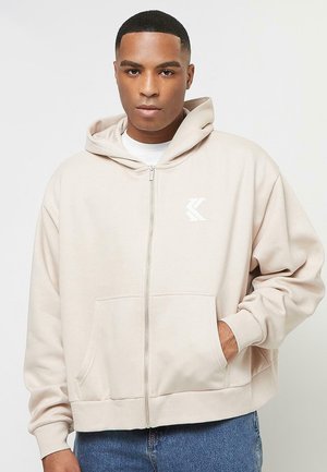 Felpa beige con zip, dotata di tasca a marsupio, polsini a costine e un logo bianco sul lato sinistro del petto. Tessuto morbido con una vestibilità comoda.