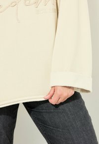 Sudadera de forro polar color crema con texto bordado "EST. 1987" en el frente. Presenta puños anchos y un corte relajado, combinada con jeans oscuros.