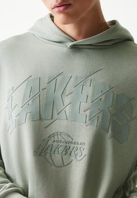 Πράσινο φούτερ με κουκούλα με ανάγλυφο, υπερμεγέθες λογότυπο «LAKERS» σε πιο σκούρα απόχρωση. Διαθέτει γραφικό μπάσκετ και μαλακό, λείο ύφασμα.