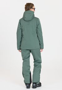 Grüne, wasserdichte Jacke mit Kapuze, taillierter Schnitt, glatte Textur, kombiniert mit passenden grünen Skihosen und schwarzen Stiefeln mit Akzenten.
