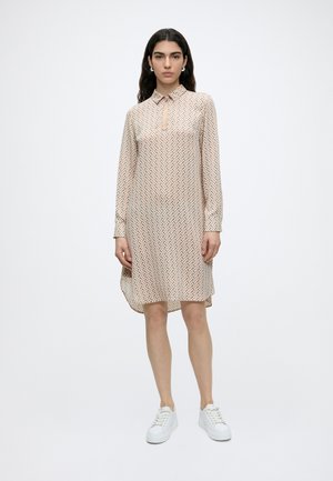 Femme aux longs cheveux foncés portant des boucles d'oreilles en perles, une robe beige à manches longues à motif avec un décolleté en trou de serrure, et des baskets blanches, debout.