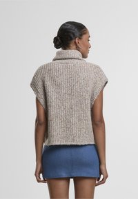 Kootud lühikeste varrukatega sviiter heledas beežis, millel on mitmevärvilised laigud ja kõrge kaelus, kantud tumesinise teksaminise seelikuga.