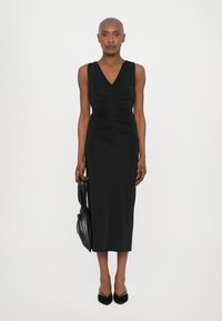 Robes midi sans manches noires avec un décolleté en V, des détails froncés à la taille et une texture lisse. Portée avec des talons aiguilles noirs et un sac à main.