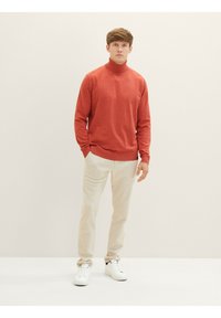 Un pull roulé rouge, texture tricotée, associé à un pantalon clair et des baskets blanches. Design simple, accents minimaux.