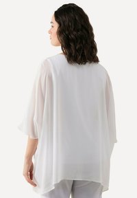 Blusa bianca, trasparente, con maniche a tre quarti e orlo curvo, che mette in evidenza un tessuto morbido e leggero e un design fluido.