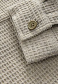 Beige textured fabric with a grid pattern; features a button closure. The surface is soft and slightly fuzzy, providing a cozy appearance.