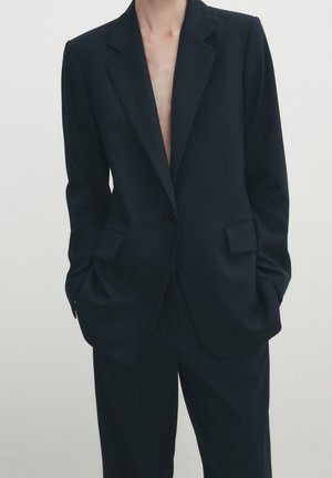 Person wearing a black tailored suit with a single-button blazer and matching trousers, hands in pockets against a plain background.