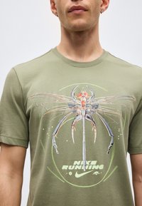 Άνδρας που φοράει ελιάς πράσινο Nike Running t-shirt με μεγάλο αφηρημένο γραφικό έντομο στο στήθος και λογότυπο Nike swoosh από κάτω.