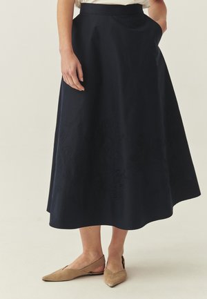 Naine, kes kannab musta midi pikkusega seelikut kerge tikandiga, käsi taskus, beežide teravate nina ja kannaga slingback kingade ja heleda pealsega.