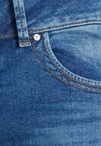Gros plan d'une poche de jean en denim bleu avec une couture orange et un rivet métallique près de la boucle de ceinture.