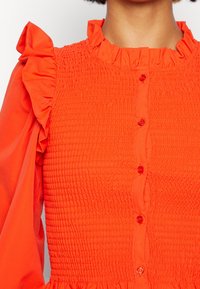 Femme portant un chemisier orange vif avec devant smocké, col à volants et détail à volants aux épaules, boutons au centre.
