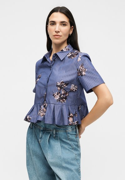 Femme portant une chemise bleu courte à manches courtes avec des motifs floraux et un ourlet peplum, associée à un jean taille haute bleu clair.