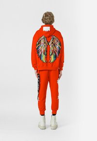 Sudadera roja con capucha y pantalones de chándal con un diseño de alas de lentejuelas multicolores en la parte posterior, combinados con botas blancas hasta el tobillo y detalles en negro en las correas.