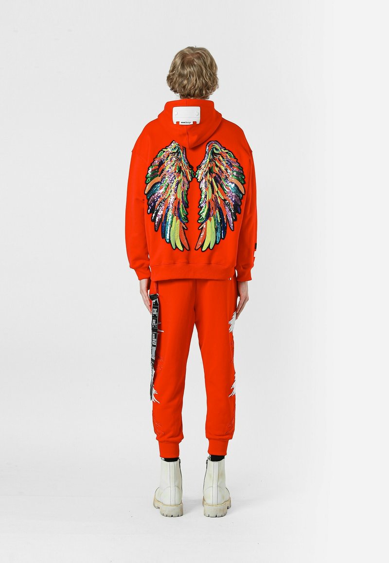 Sudadera roja con capucha y pantalones de chándal con un diseño de alas de lentejuelas multicolores en la parte posterior, combinados con botas blancas hasta el tobillo y detalles en negro en las correas.