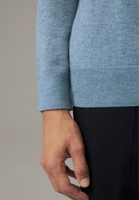 Cuff di un maglione a maglia azzurro chiaro con texture a coste, abbinato a un tessuto scuro sottostante. La mano mostra un tono di pelle naturale e unghie.
