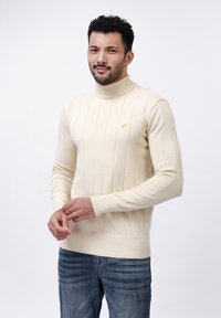 Beige bordás garbós pulóver, testhez simuló fazonnal és hosszú ujjakkal, a mellrészen egy kis sárga logóval. Kék farmernadrággal viselve.