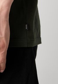 Mørkegrøn bomuld t-shirt med en side søm label. Har en glat tekstur, korte ærmer og en lige kant med synlige sting.