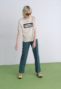Beige grafisk t-skjorte med svart "JORDAN" logo, kombinert med mørkegrønne rett-leggete bukser. Modellen har på seg brune sneakers og solbriller.