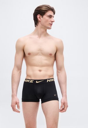 Άνδρας χωρίς πουκάμισο με κοντά καστανά μαλλιά, φορώντας μαύρα εσώρουχα τύπου μπόξερ Nike με χρυσό λογότυπο στη λαστιχένια μέση, κοιτάει προς τα αριστερά του.