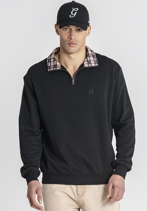 Man draagt zwarte quarter-zip trui met geruite kraag, beige broek en zwarte baseballpet met witte letter G voorop.