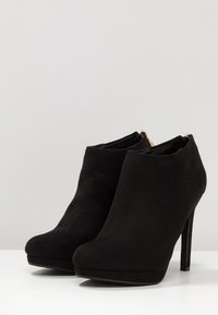 Černé semišové kotníkové boty s hladkým povrchem, špičatou špičkou a tenkým jehlovým podpatkem. Obsahují zadní zip pro zapínání.