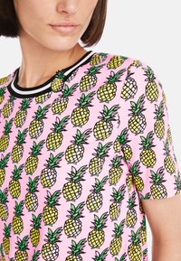 Kurzarmhemd mit einem pinken Hintergrund und einem sich wiederholenden Ananasdruck in Gelb und Grün. Der gerippte schwarze Kragen sorgt für einen Kontrast.