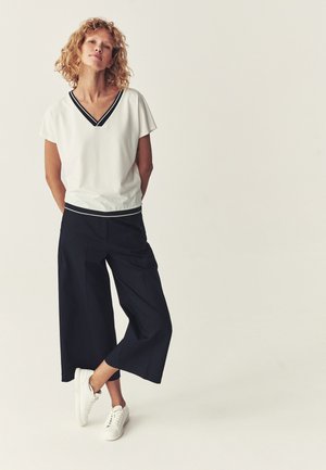 Nő áll, fehér V-nyakú rövid ujjú felsőt, fekete széles szárú, rövidített nadrágot és fehér sportcipőt visel egyszínű háttér előtt.